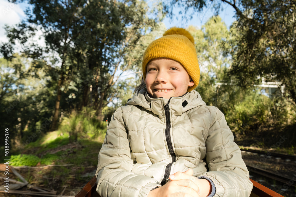 Portrait of happy boy in yellow hat and grey down jacket enjoying a ride on miniature train in the park.