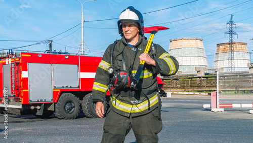 A lifeguard in protective clothing and helmet holds an axe on his shoulder. A firefighter next to a fire truck on the background of factory pipes. Rescue service. Rescue of people in case of fires.