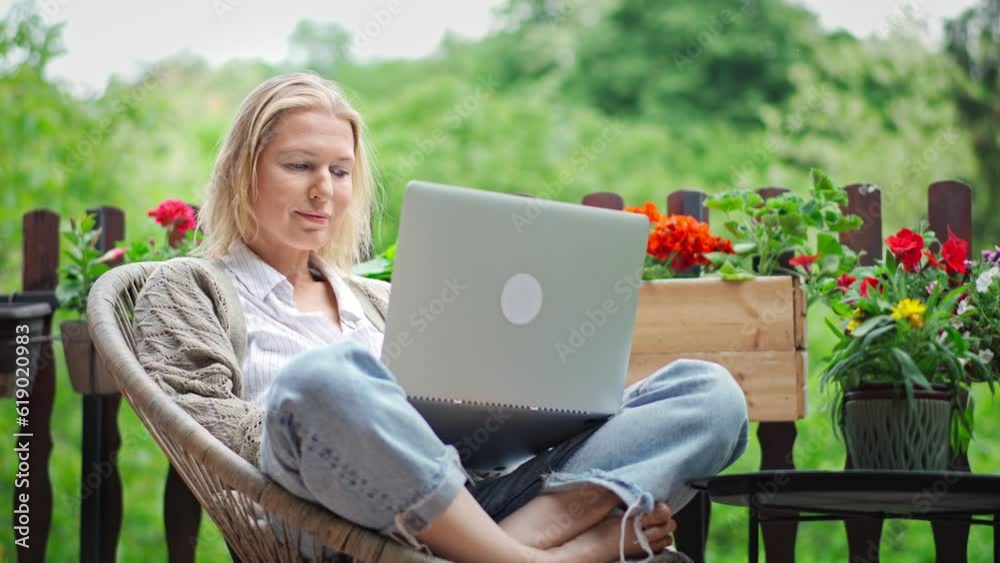 A middle-aged Caucasian woman working on a laptop while sitting on the terrace of a village house. A middle-aged Caucasian woman working on a laptop while sitting on the terrace of a village house.