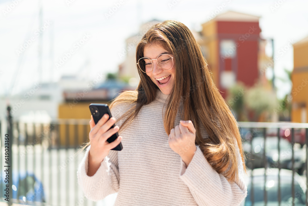 Young pretty caucasian woman with glasses at outdoors with phone in victory position