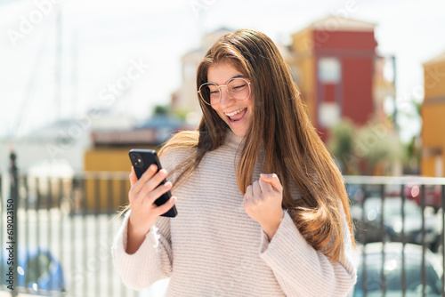 Young pretty caucasian woman with glasses at outdoors with phone in victory position