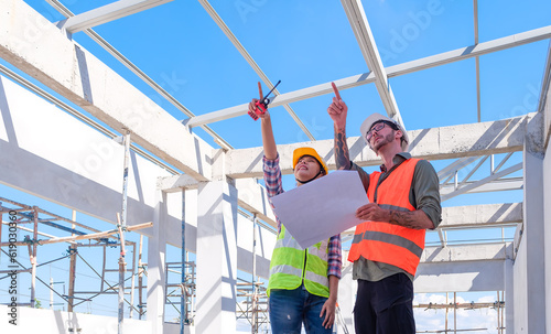 Caucasian man and Asian woman engineers in protective clothes and hardhat inspecting the construction work on site with blueprint. Cooperation concept.