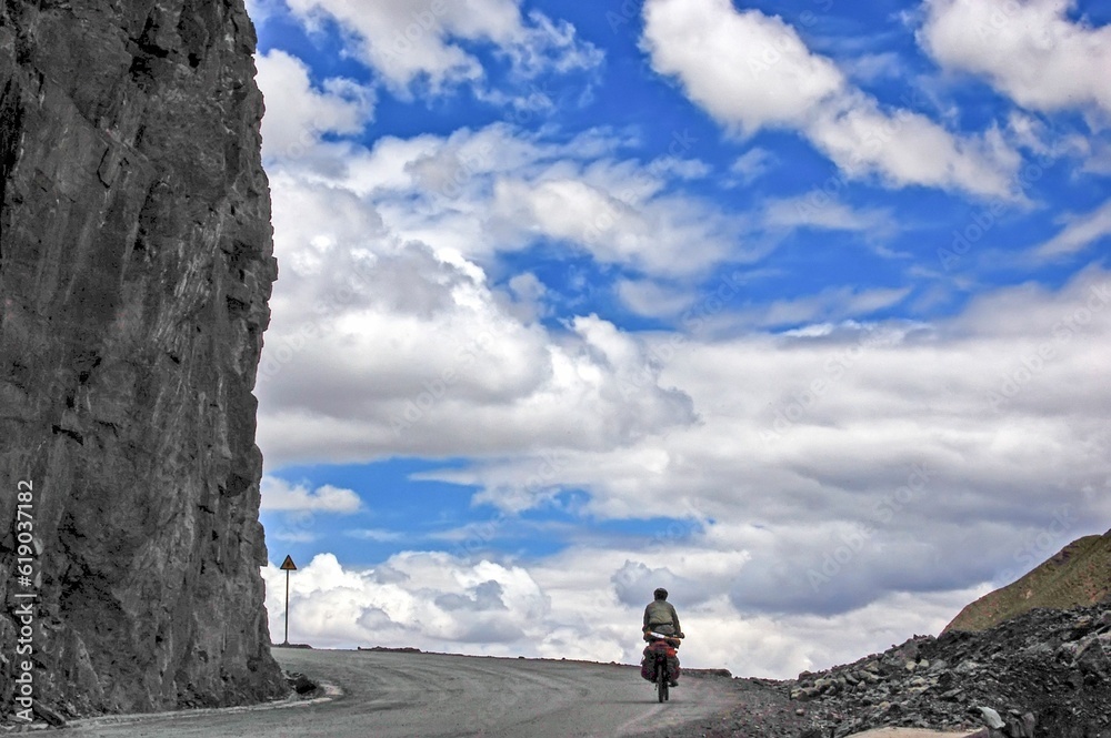 Cyclist riding on the empty road next to a huge cliff with the blue cloudy sky in the background