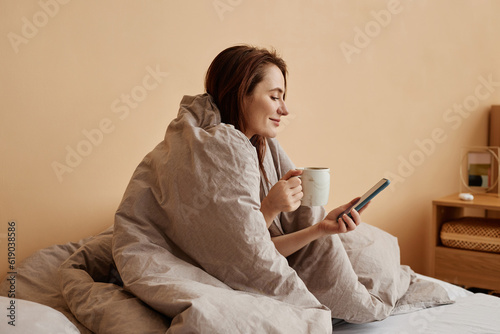 Minimal side view portrait of young woman covered in cozy blanket enjoying relaxing weekend at home and holding phone, copy space