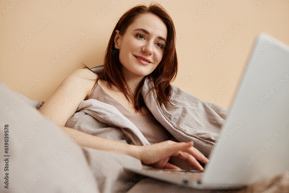 Cozy portrait of young woman using laptop computer in bed and smiling, copy space Cozy portrait of young woman using laptop computer in bed and smiling, copy space