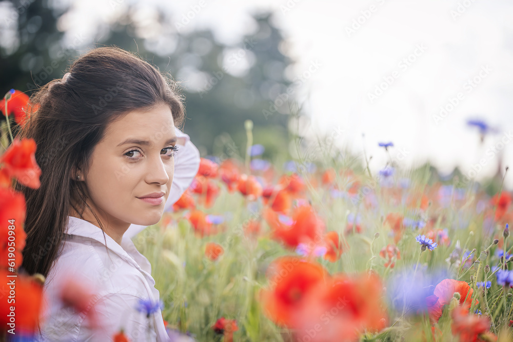 Naklejka premium Side view of atractive brunette girl with hand in her hair posing in a red poppies field. Horizontally. 