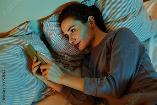 Top view portrait of smiling adult woman using smartphone in bed at night reading text messages and scrolling social media photo
