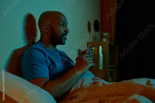 Side view portrait of smiling black man watching TV in bed at night staying up late, copy space
