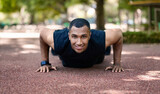 Smiling handsome millennial black sportsman doing push ups on track at summer park, at morning, panorama