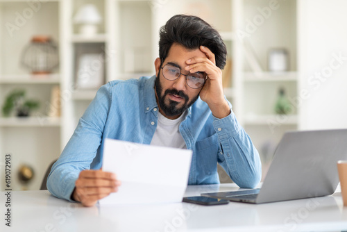 Upset Young Indian Man Sitting At Desk With Laptop And Checking Documents
