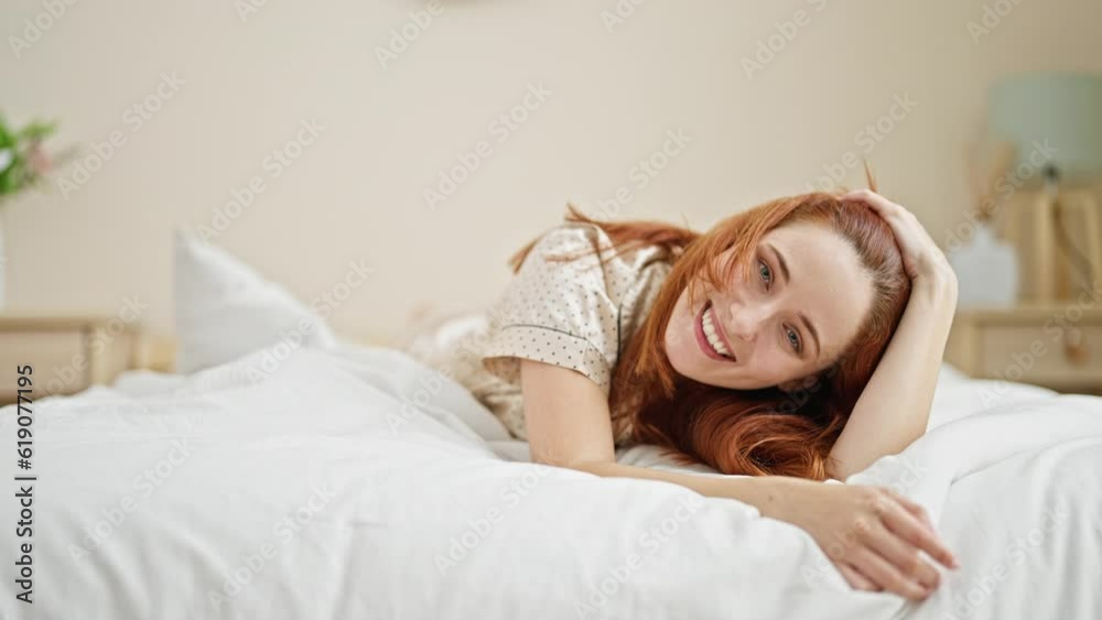 Young redhead woman touching hair lying on bed smiling at bedroom