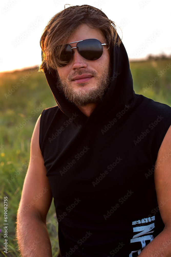 a young blond guy in a black hooded T-shirt and sun goggles is sitting on the grass enjoying the rays of the setting sun a young blond guy in a black hooded T-shirt and sun goggles is sitting on the grass enjoying the rays of the setting sun
