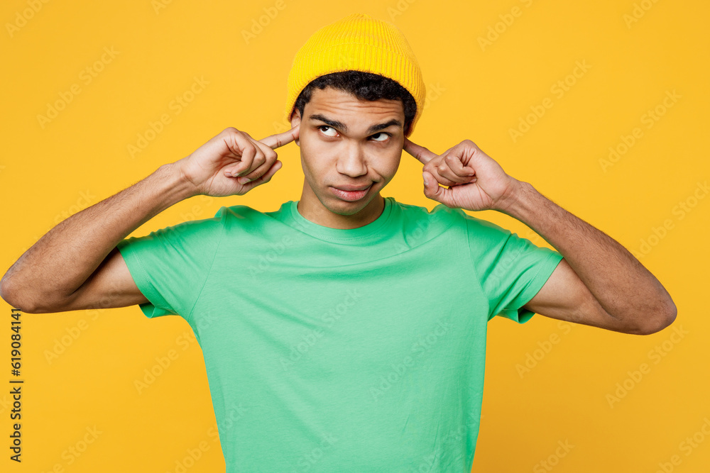 Young sad man of African American ethnicity he wearing casual clothes green t-shirt hat cover ears with hands fingers do not want to listen scream isolated on plain yellow background studio portrait. Young sad man of African American ethnicity he wearing casual clothes green t-shirt hat cover ears with hands fingers do not want to listen scream isolated on plain yellow background studio portrait.