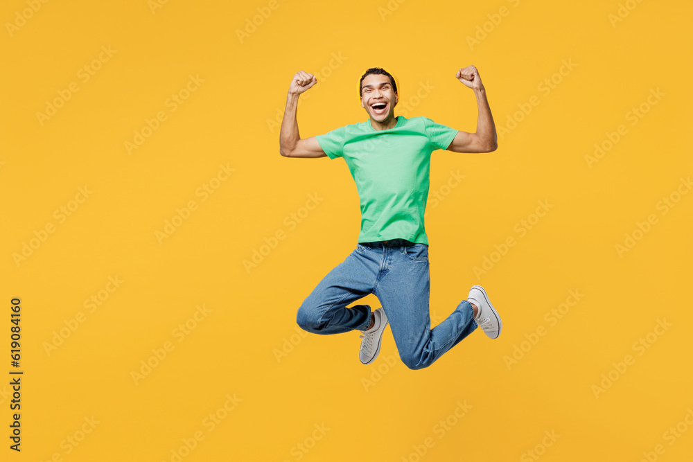 Full body young man of African American ethnicity he wears casual clothes green t-shirt hat jump high doing winner gesture celebrate clenching fists say yes isolated on plain yellow background studio.