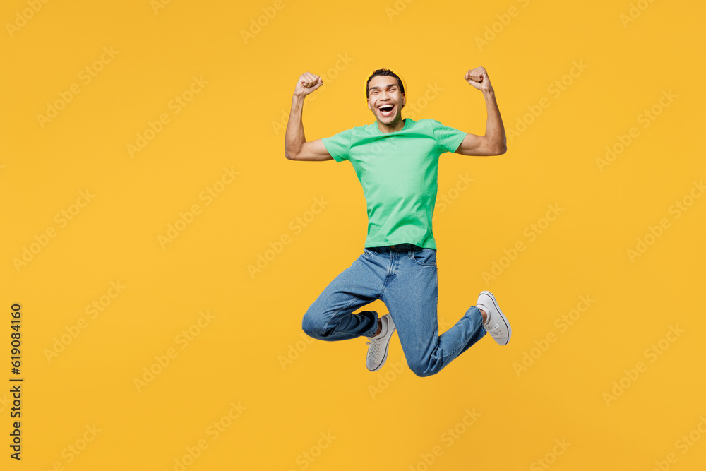 Full body young man of African American ethnicity he wears casual clothes green t-shirt hat jump high doing winner gesture celebrate clenching fists say yes isolated on plain yellow background studio. Full body young man of African American ethnicity he wears casual clothes green t-shirt hat jump high doing winner gesture celebrate clenching fists say yes isolated on plain yellow background studio.