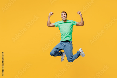 Full body young man of African American ethnicity he wears casual clothes green t-shirt hat jump high doing winner gesture celebrate clenching fists say yes isolated on plain yellow background studio. © ViDi Studio
