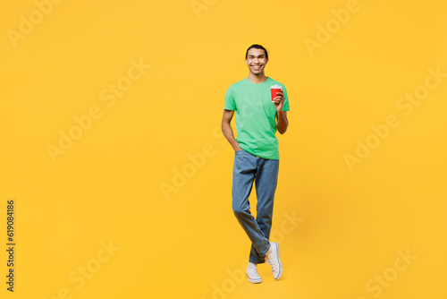 Full body happy young man of African American ethnicity he wears casual clothes green t-shirt hat hold takeaway delivery craft paper brown cup coffee to go isolated on plain yellow background studio.