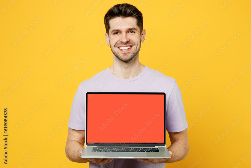 Young smiling cheerful happy IT man wears light purple t-shirt casual clothes hold use work on laptop pc computer with blank screen workspace area isolated on plain yellow background studio portrait.