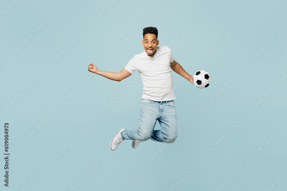 Fototapeta premium Full body overjoyed young man fan wears t-shirt do winner gesture cheer up support football sport team hold soccer ball jump high watch tv live stream isolated on plain pastel blue color background.