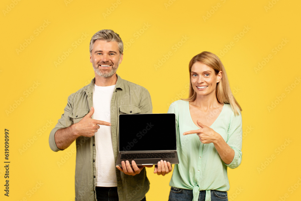 Happy european middle aged couple pointing fingers at laptop with blank black screen, yellow background, mockup Happy european middle aged couple pointing fingers at laptop with blank black screen, yellow background, mockup