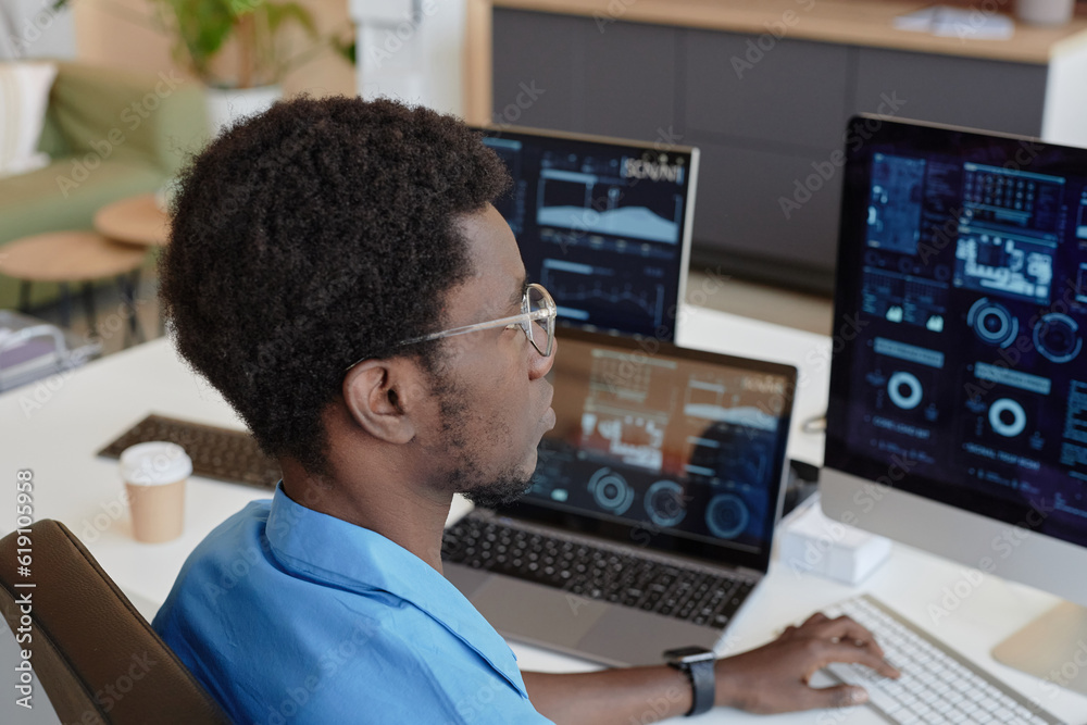 Young black man in eyeglasses looking at graphs and charts on computer screens while sitting by workplace in office and analyzing data