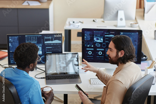 Young confident IT engineer explaining graphic data on computer screens to his colleague while looking at him during working meeting
