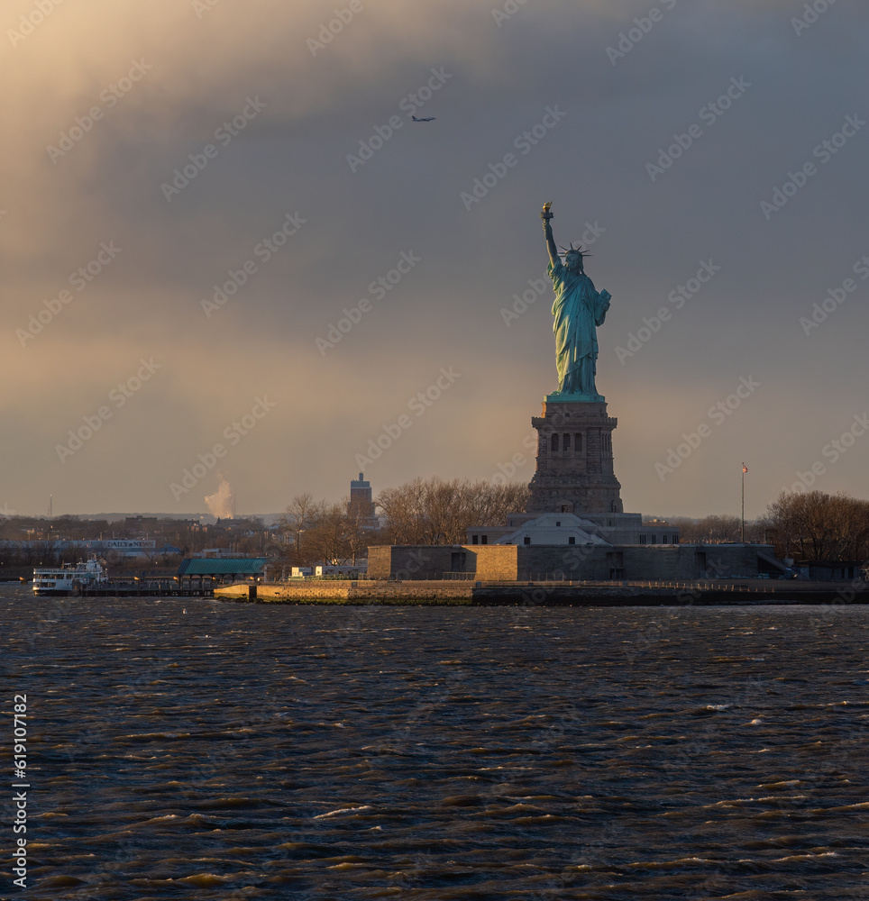 View of the Statue of Liberty during the day with boat docked on the island and Plane flying overhead