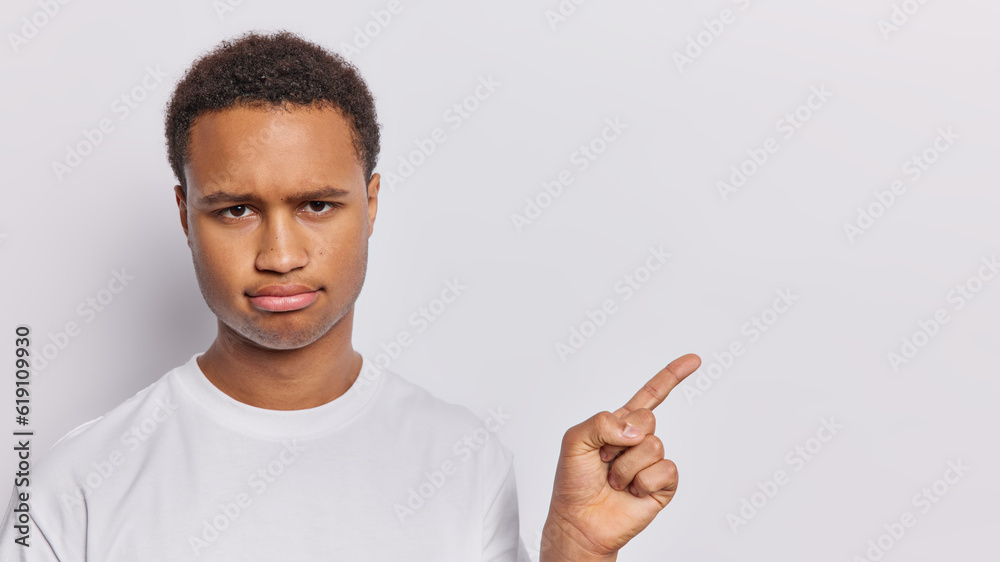 Sad young curly haired man pointing with index finger backwards has discontent expression tells about something unpleasant dressed in casual t shirt isolated over white background. Look at this Sad young curly haired man pointing with index finger backwards has discontent expression tells about something unpleasant dressed in casual t shirt isolated over white background. Look at this
