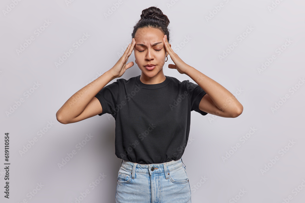 Beautiful Latin woman suffers from headache keeps hands on temples feels exhausted tries to concentrate wears casual black t shirt and jeans isolated over white background. Health problems concept Beautiful Latin woman suffers from headache keeps hands on temples feels exhausted tries to concentrate wears casual black t shirt and jeans isolated over white background. Health problems concept