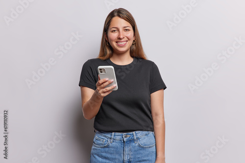 Horizontal shot of cheerful young woman with long straight hair holds mobile phone for chatting online connected to wireless internet wears black t shirt and jeans isolated over white background