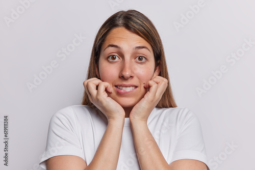 Nervous worried long haired European woman bites lips keeps hands on cheeks worries before important event dressed in casual t shirt isolated over white background hears unexpected bad news.