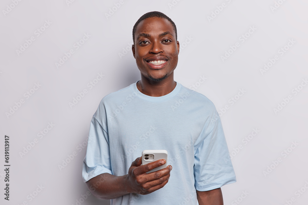 Photo of black bearded man uses modern mobile phone for chatting online and browsing internet being always in touch smiles toothily dressed in casual blue t shirt stands against white background Photo of black bearded man uses modern mobile phone for chatting online and browsing internet being always in touch smiles toothily dressed in casual blue t shirt stands against white background
