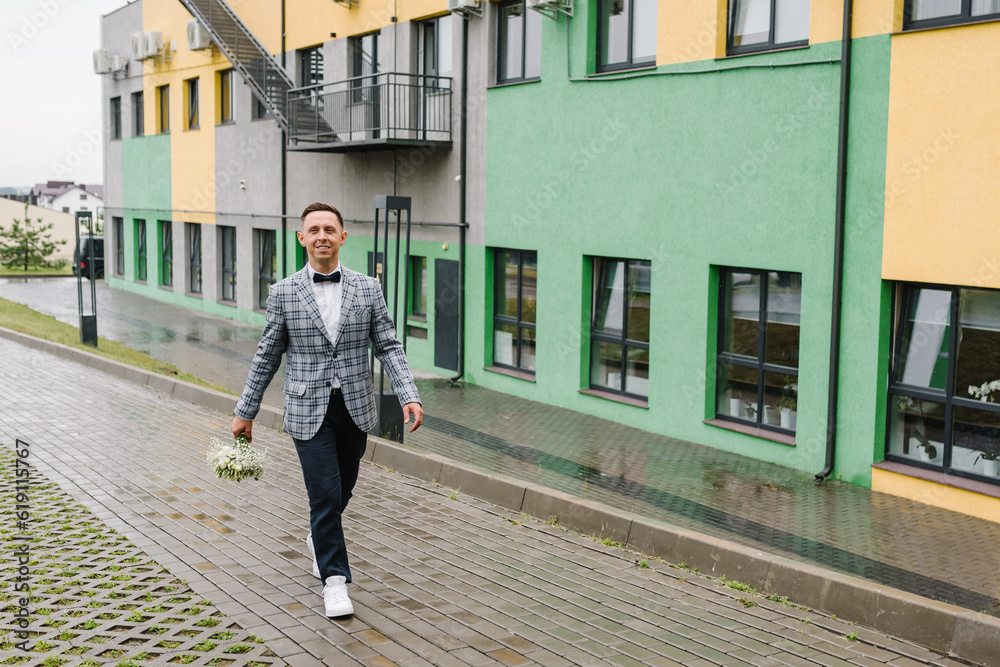 Handsome happy groom with bouquet walking in the near house. Portrait of the man. Wedding morning on a rainy day.