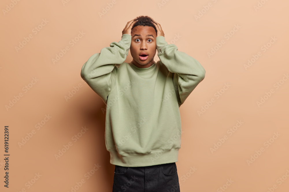 Terrified shocked black young man keeps hands on head has widely opened mouth cannot believe in something impressive dressed in casual jumper ans black trousers isolated over brown background. Terrified shocked black young man keeps hands on head has widely opened mouth cannot believe in something impressive dressed in casual jumper ans black trousers isolated over brown background.