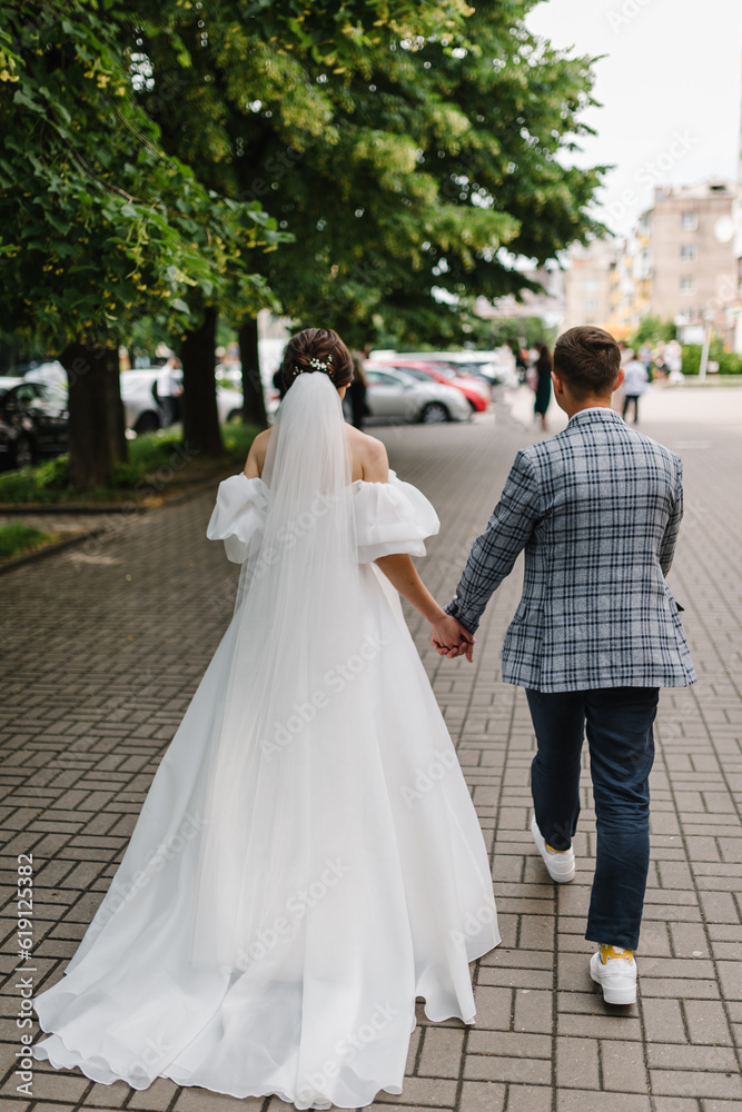 A young, stylish groom and beautiful bride in a white dress walking in the alley in the city against the background of buildings, trees. Wedding photography of the newlyweds on the street. Back view.