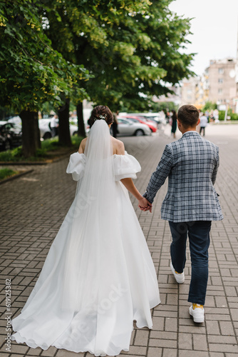 A young, stylish groom and beautiful bride in a white dress walking in the alley in the city against the background of buildings, trees. Wedding photography of the newlyweds on the street. Back view. © Serhii