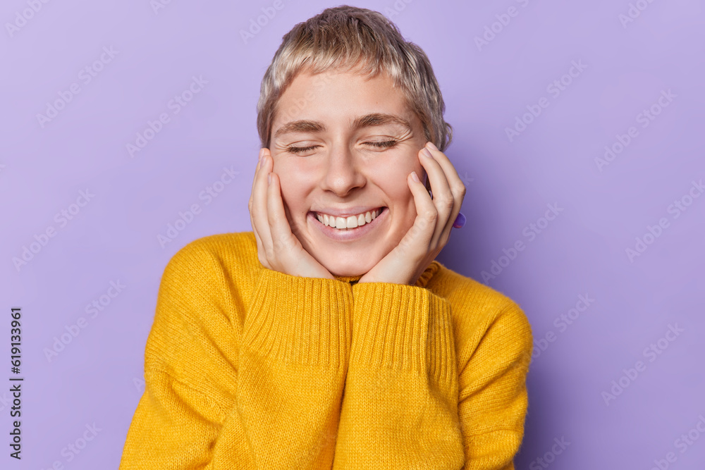Portrait of cheerful woman with blonde short hair keeps eyes closed touches cheeks smiles gently expresses happy sincere emotions wears yellow jumper isolated over purple wall. People and positiveness Portrait of cheerful woman with blonde short hair keeps eyes closed touches cheeks smiles gently expresses happy sincere emotions wears yellow jumper isolated over purple wall. People and positiveness