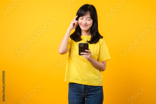 Experience range of emotions with expressive portrait. young Asian woman wearing yellow t-shirt and denim jeans showcases doubtful expression while using smartphone. diverse emotions and tech usage.