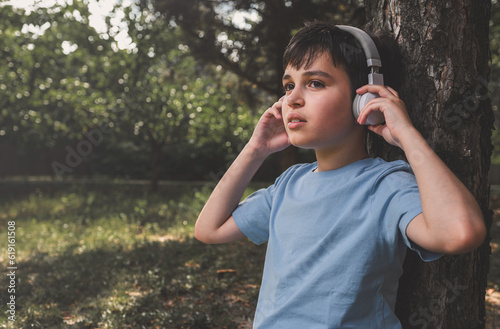 Toned portrait of adorable dark haired teenage boy listening to music on wireless headphones sitting under tree in park