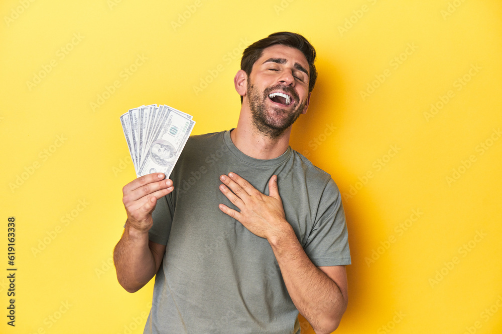 Caucasian man holding dollars, yellow studio shot laughs out loudly keeping hand on chest. Caucasian man holding dollars, yellow studio shot laughs out loudly keeping hand on chest.