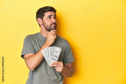 Photographie Caucasian man holding dollars, yellow studio shot looking sideways with doubtful and skeptical expression