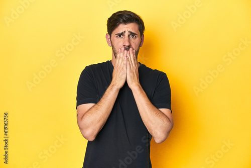 Caucasian man in black t-shirt, yellow studio backdrop shocked, covering mouth with hands, anxious to discover something new.