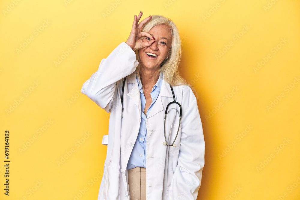 Middle-aged Caucasian female doctor on yellow studio backdrop Middle-aged Caucasian female doctor on yellow studio backdropexcited keeping ok gesture on eye. Middle-aged Caucasian female doctor on yellow studio backdrop Middle-aged Caucasian female doctor on yellow studio backdropexcited keeping ok gesture on eye.