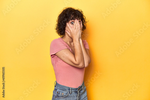 Curly-haired Caucasian woman in pink t-shirt blink through fingers frightened and nervous. © Asier