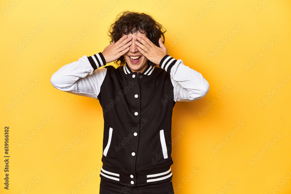 Curly-haired Caucasian woman in baseball jacket covers eyes with hands, smiles broadly waiting for a surprise. Curly-haired Caucasian woman in baseball jacket covers eyes with hands, smiles broadly waiting for a surprise.