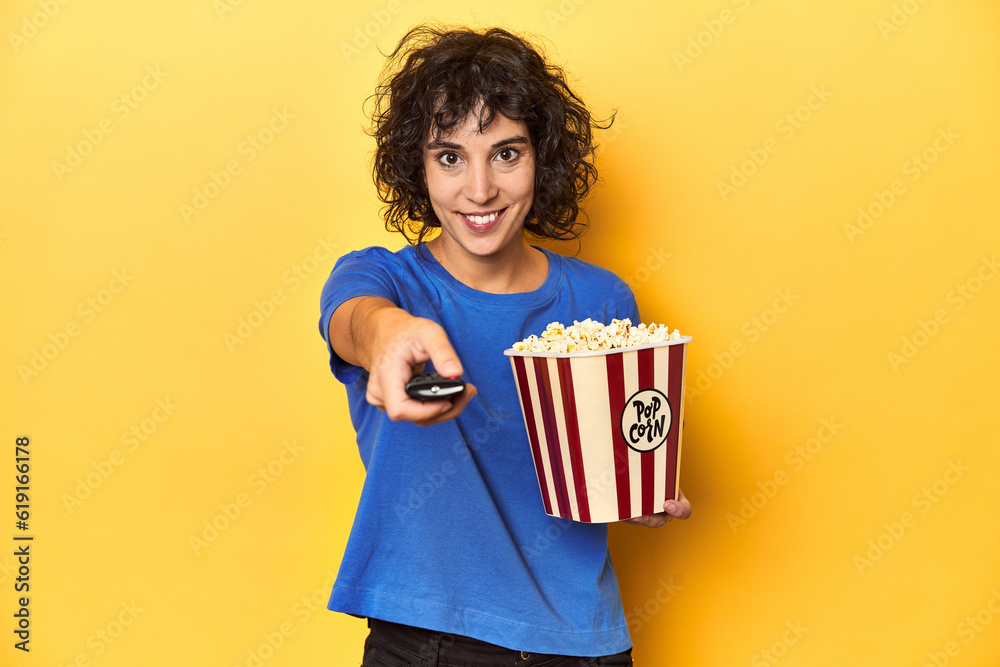 Woman with popcorn and TV remote, home cinema concept, yellow studio backdrop.