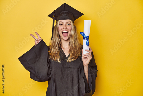 Young caucasian woman wearing a graduation robe holding a diploma Yjoyful and carefree showing a peace symbol with fingers.