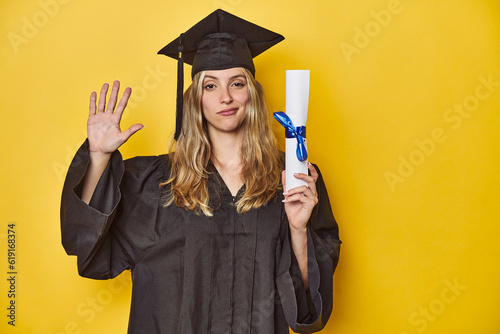 Young caucasian woman wearing a graduation robe holding a diploma Ysmiling cheerful showing number five with fingers. photo