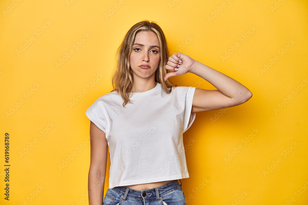Young blonde Caucasian woman in a white t-shirt on a yellow studio background, showing a dislike gesture, thumbs down. Disagreement concept. Young blonde Caucasian woman in a white t-shirt on a yellow studio background, showing a dislike gesture, thumbs down. Disagreement concept.