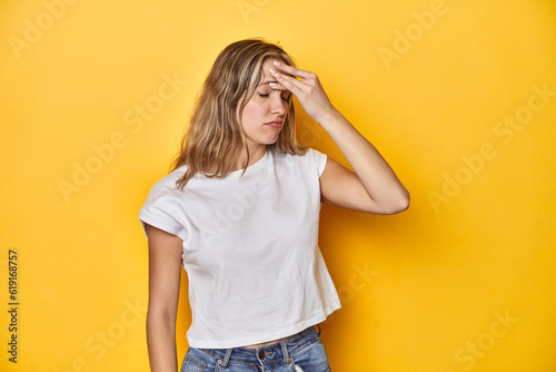 Young blonde Caucasian woman in a white t-shirt on a yellow studio background, having a head ache, touching front of the face.